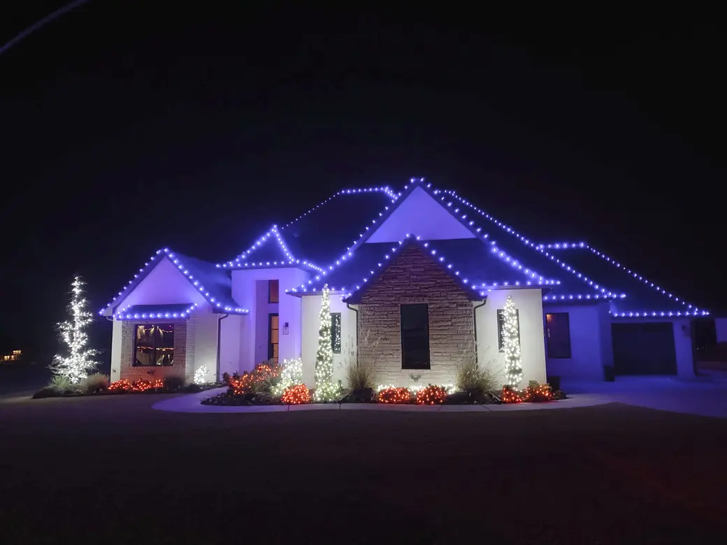 A modern stone and render home with deep blue bulbs along the rooflines, warm white cone trees and red-lit shrubs in the beds.