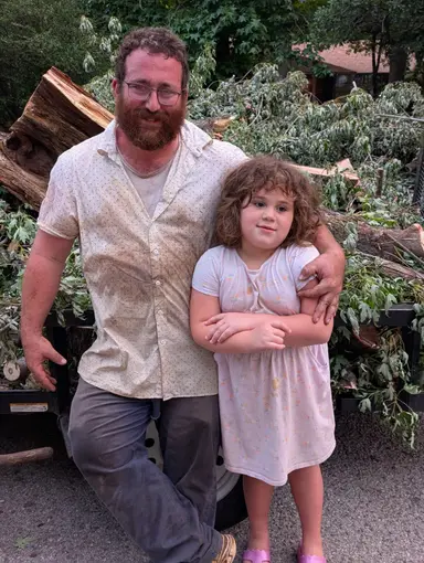 Justin Marks and his daughter beside a trailer loaded with cut timber after clearing a storm-felled tree.
